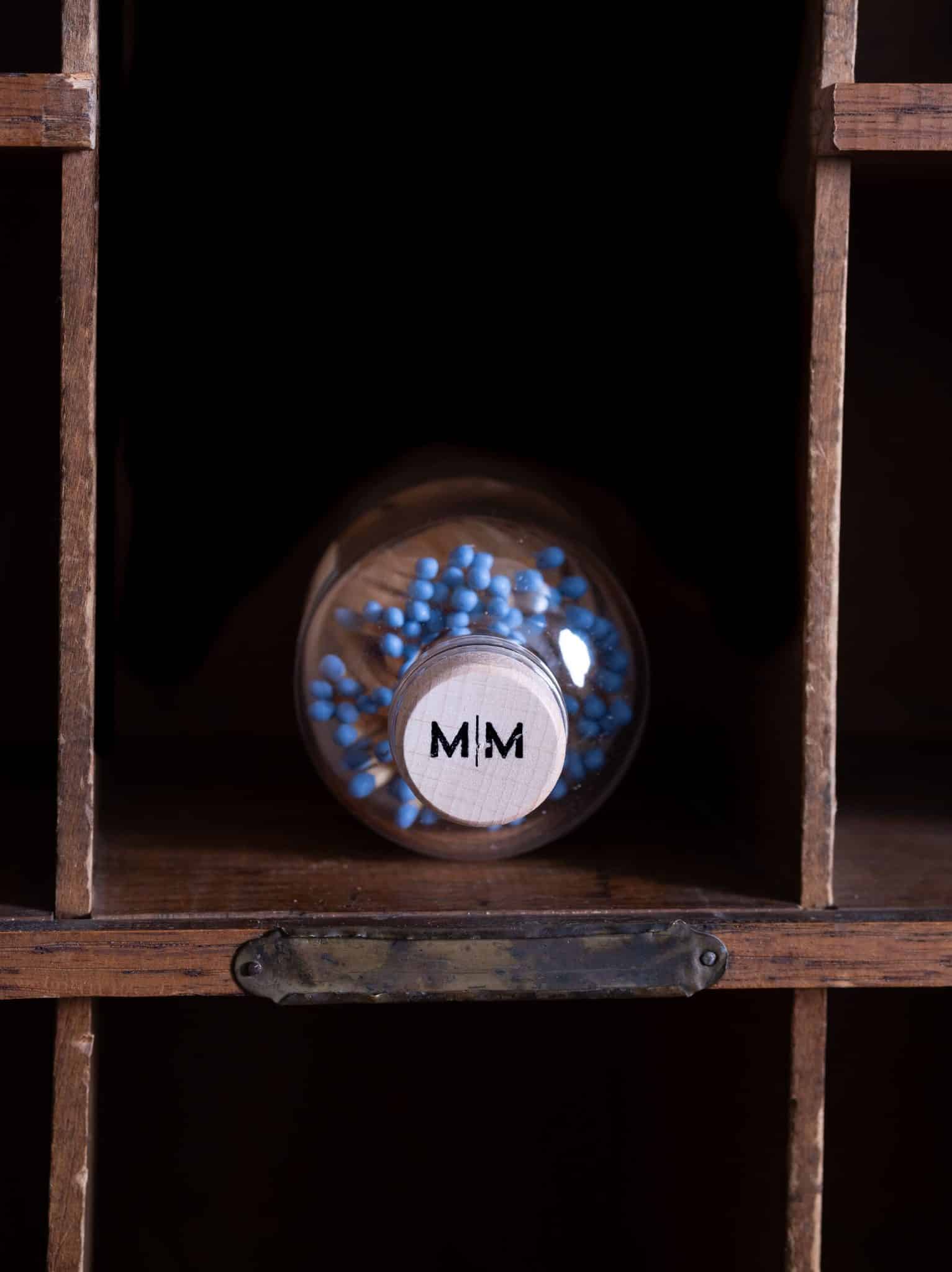 Vintage Apothecary Blue Matches in a glass jar with a cork top, with blue match tips, displayed on a wooden shelf. The label says 'Wooden Safety Matches' in black and white.