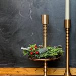 Brass Pedestal Small styled with a greenery Christmas Candle Wreath, shown beside the Taper Holder Brass Set Assorted with a white taper candle.