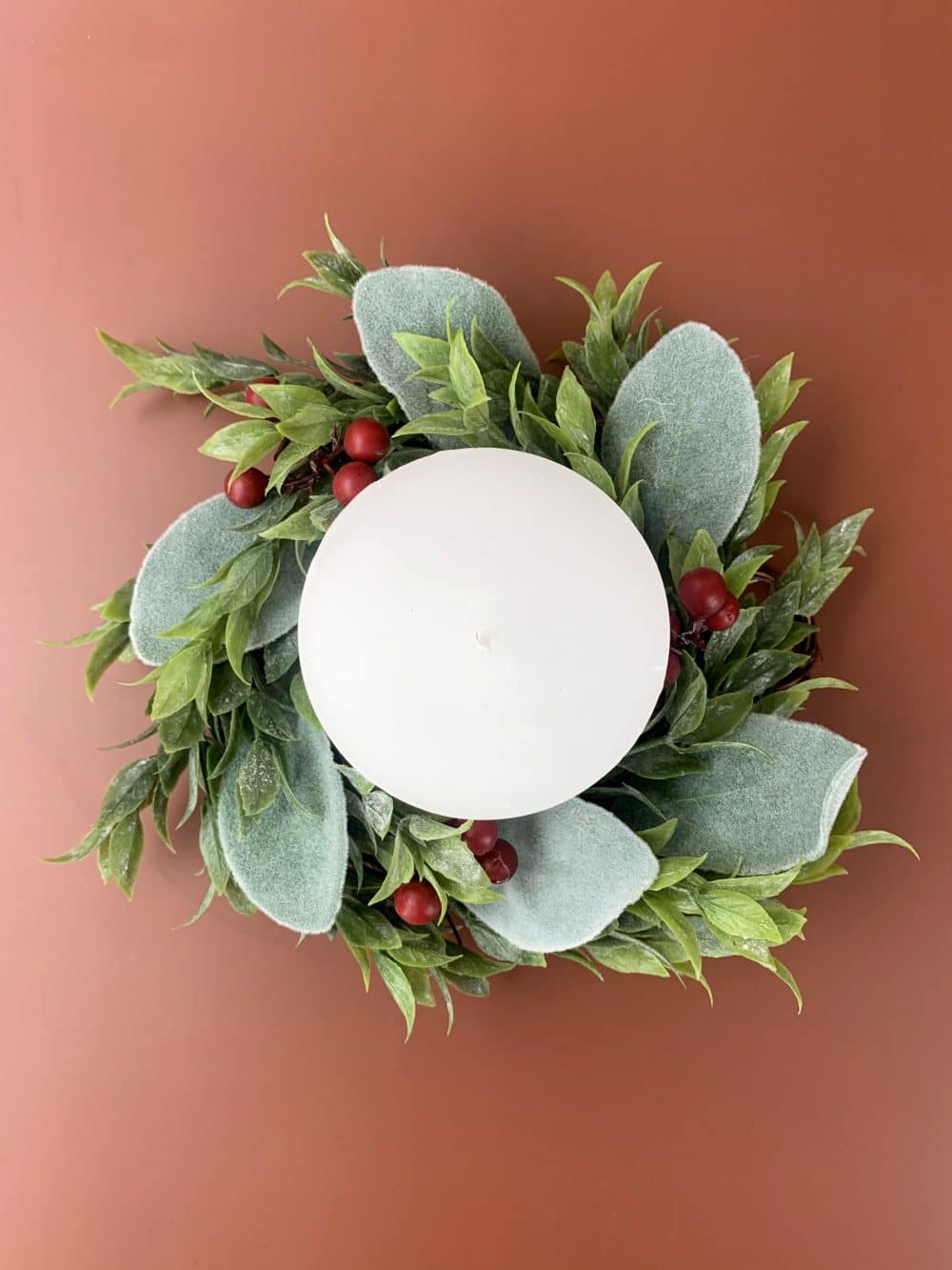 Holiday wreath with green leaves and red berries wrapped around a white candle, viewed from above.
