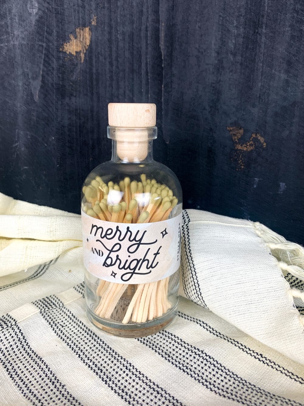 Glass apothecary jar filled with gold-tipped matches, featuring a "Merry & Bright" holiday label and wooden cork top.