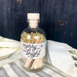 Glass apothecary jar filled with gold-tipped matches, featuring a "Merry & Bright" holiday label and wooden cork top.