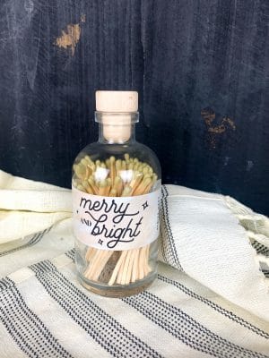 Glass apothecary jar filled with gold-tipped matches, featuring a "Merry & Bright" holiday label and wooden cork top.