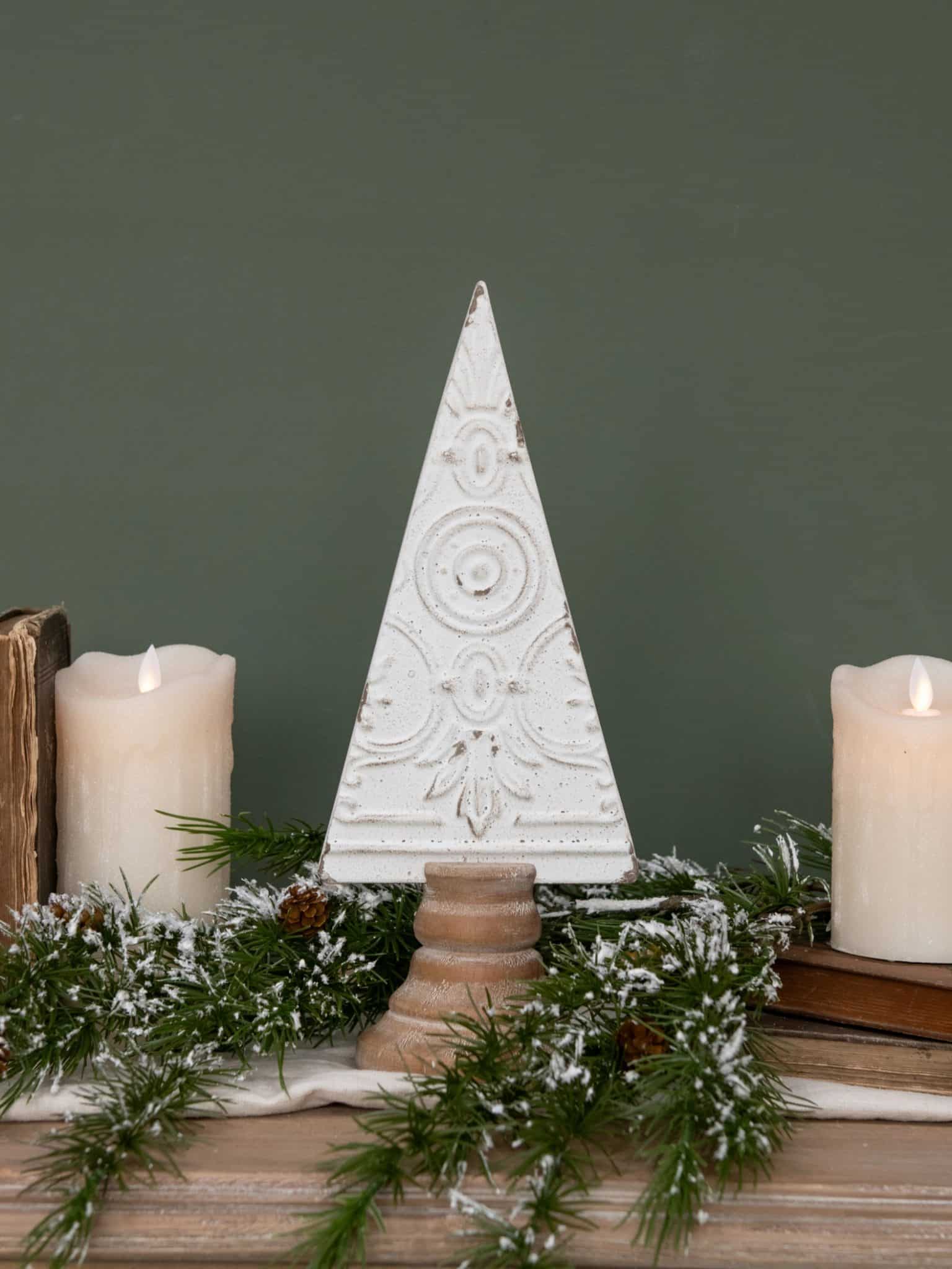 Small antique white pressed metal Christmas tree with ornate embossed design and natural wood base, styled with candles and greenery.