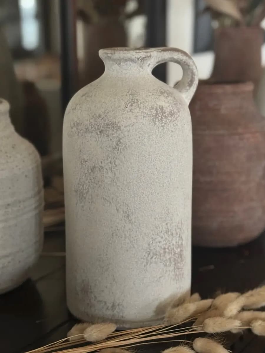 Tall rustic white vase with a single jug-style handle and textured weathered finish, styled with neutral decor.