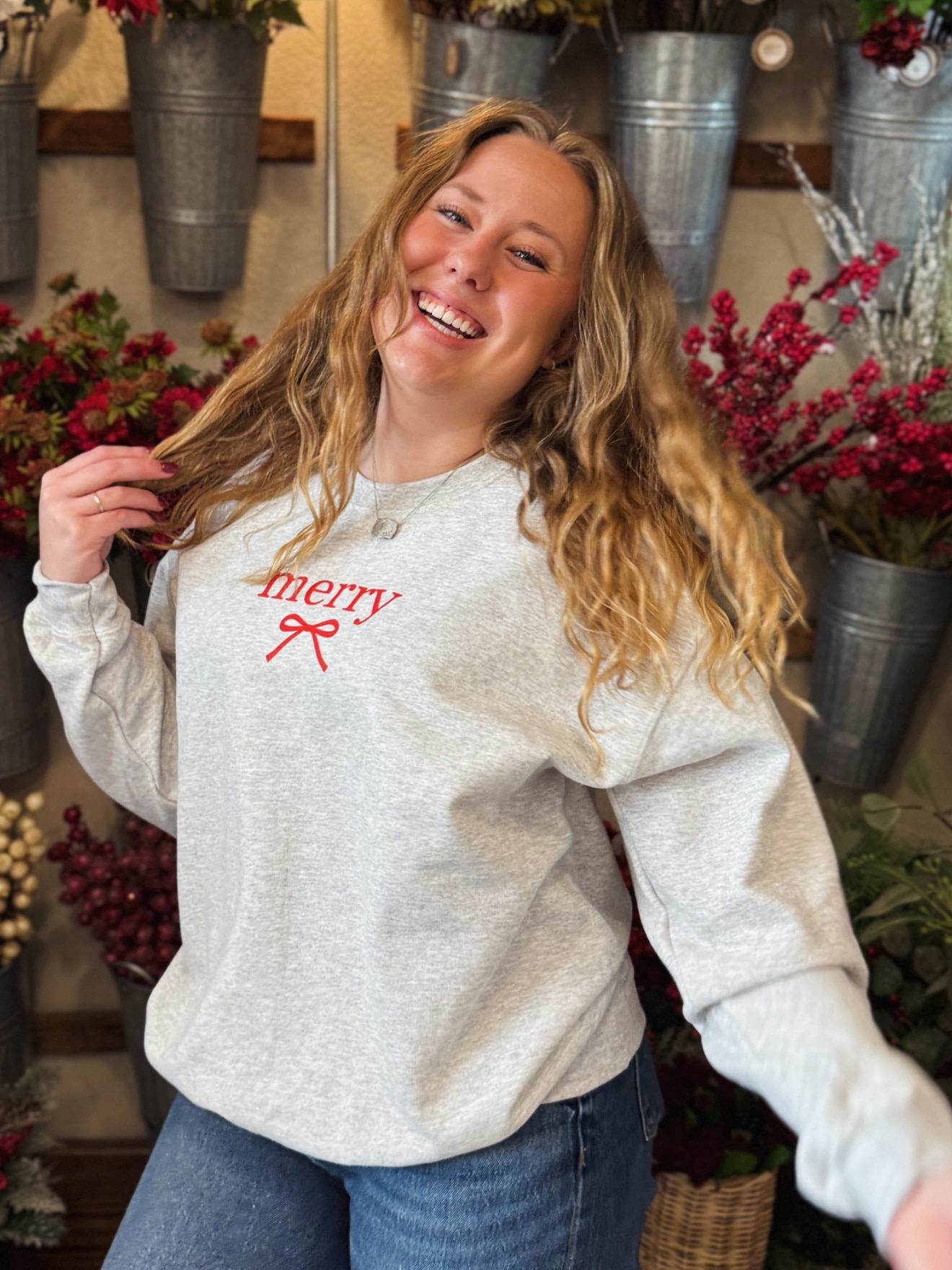 Woman wearing an ash gray crewneck with a red “merry” bow graphic, smiling in front of holiday floral buckets.