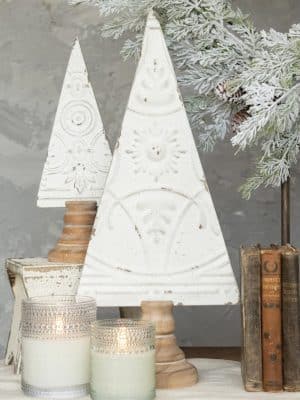 Antique white pressed metal Christmas trees with embossed detailing and wood bases, styled together with frosted greenery, books, and candles.