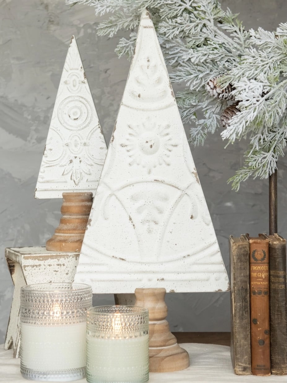 be-made-hays-ks-antique-white-pressed-metal-trees-display Antique white pressed metal Christmas trees with embossed detailing and wood bases, styled together with frosted greenery, books, and candles.