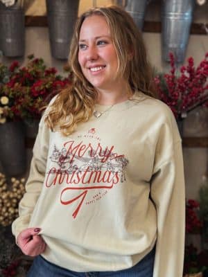 Sand crewneck with red “Merry Christmas” text and vintage Santa sleigh graphic, styled on model in front of holiday florals.