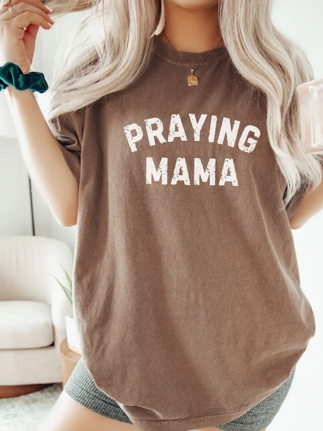 Woman wearing an espresso brown “Praying Mama” graphic tee with bold white lettering, styled casually in a cozy home setting.