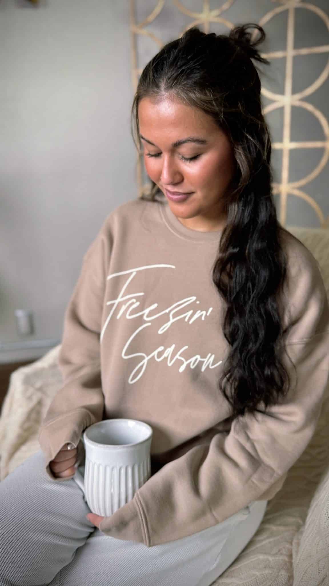 Woman wearing tan Freezin’ Season crewneck sweatshirt holding a coffee mug