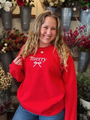 Woman wearing a Christmas red crewneck with white “merry” text and bow graphic, smiling in front of holiday florals.