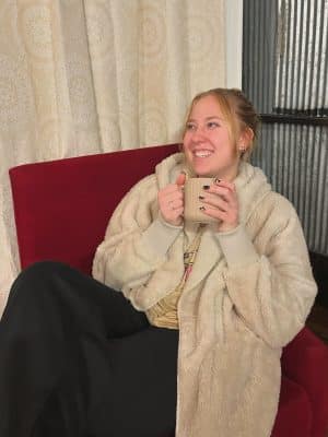 Smiling model in Taupe Plush Fur Hooded Jacket, seated in a red chair with a warm drink.