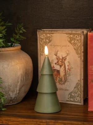 Dark green tree-shaped flameless candle styled beside a decorative book and pottery on a cozy holiday shelf.