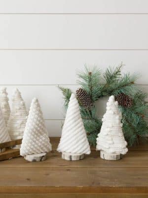 Three plush white decorative Christmas trees with textured fabric patterns and faux wood bases, styled with a pine wreath on a wooden table.