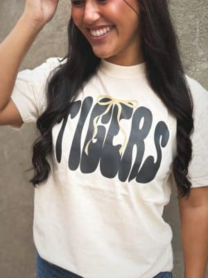 Person wearing Tigers graphic tee in ivory with oversized black lettering and gold bow design.