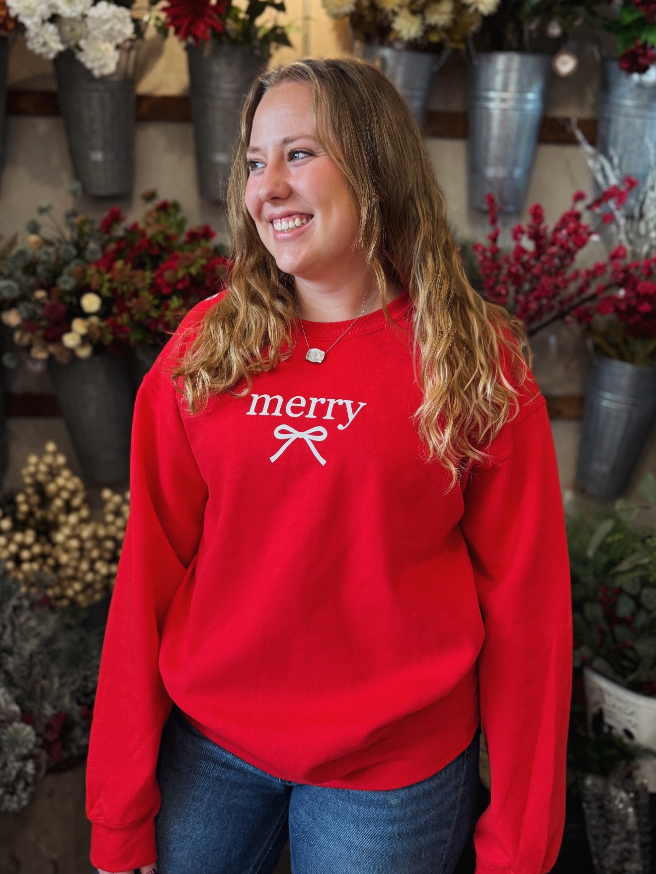 Ash-blonde woman in a red Merry Bow sweatshirt, standing in a floral shop filled with winter greenery and berries.