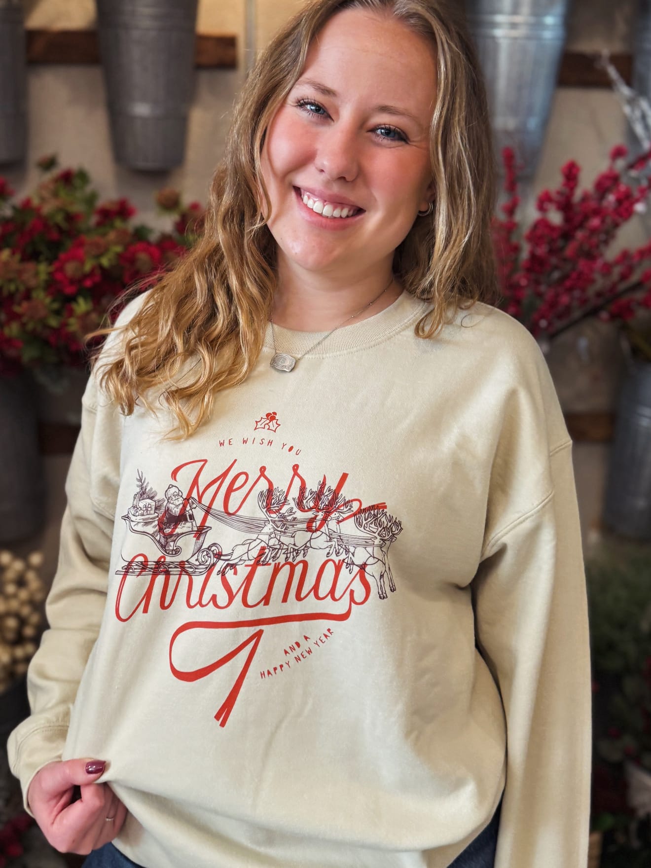 Model wearing sand-colored Merry Christmas sweatshirt featuring red script text and illustrated Santa with reindeer.