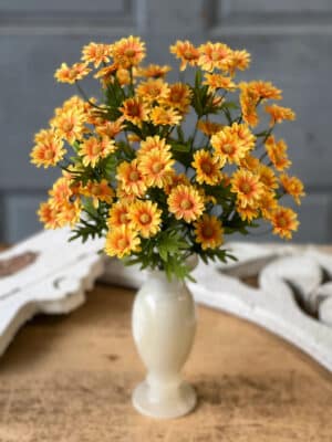 Golden Marigold Daisy Bush arranged in a cream vase with cheerful yellow-orange blooms and scalloped greenery.