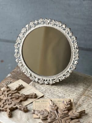 Round distressed floral mirror with ornate white frame styled on vintage papers and neutral decor.