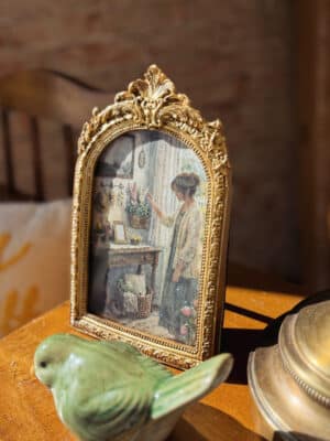 Gold ornate arched photo frame styled with a green stoneware bird figurine on a tabletop.