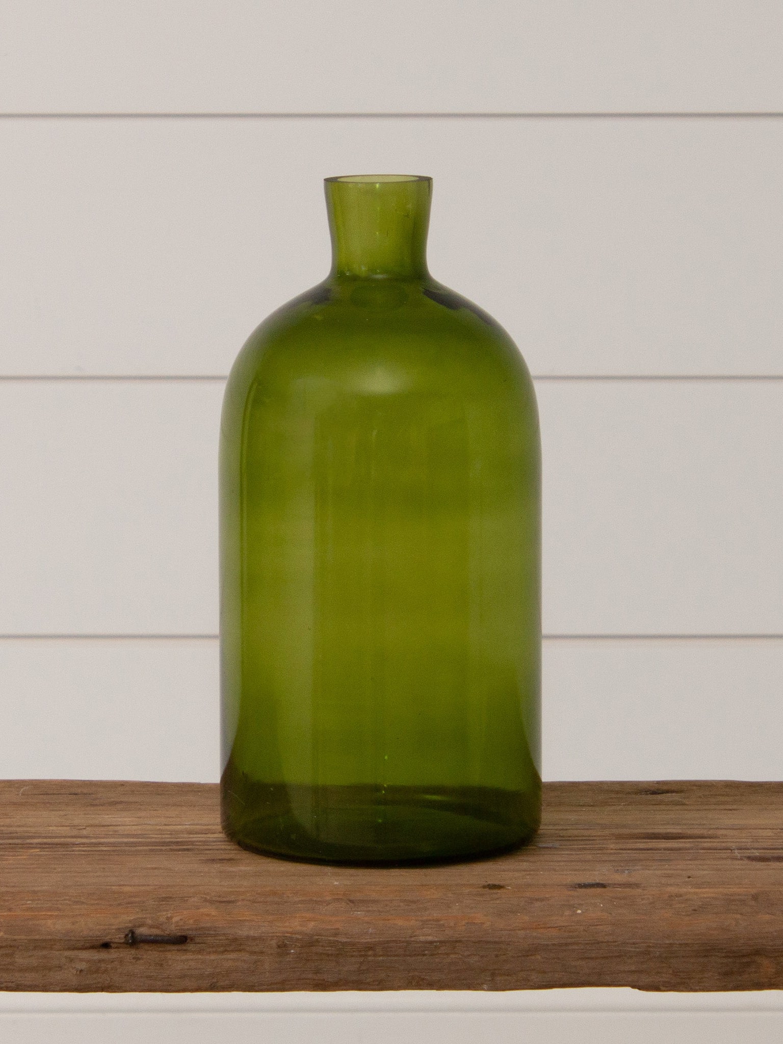 Simple green glass vase with a smooth bottle shape displayed on a rustic wood surface against a white shiplap background.