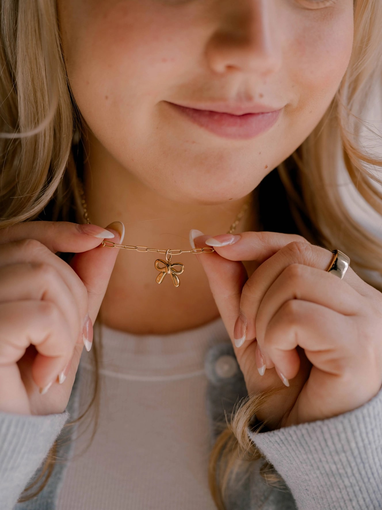 Woman wearing the He Holds All Together Charm Necklace, gently holding the gold bow pendant near her collarbone.