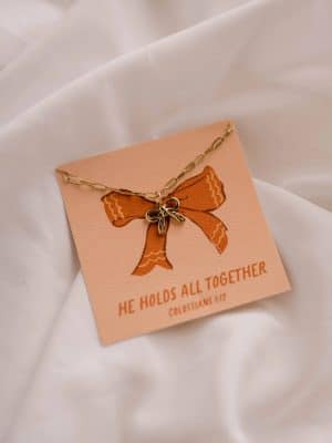 He Holds All Together Charm Necklace draped across its illustrated Scripture card on a soft white fabric background.