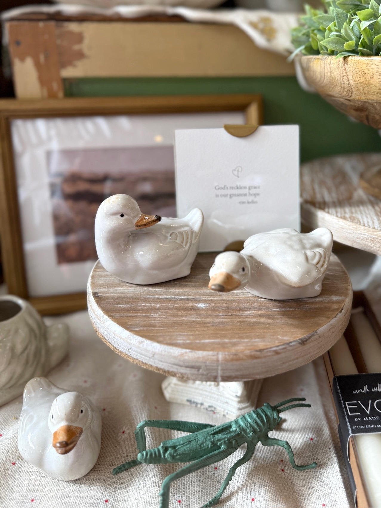 Three white stoneware vintage duckling figurines displayed on a wood and metal white cast pedestal with a green pewter grasshopper sculpture and framed decor in the background.