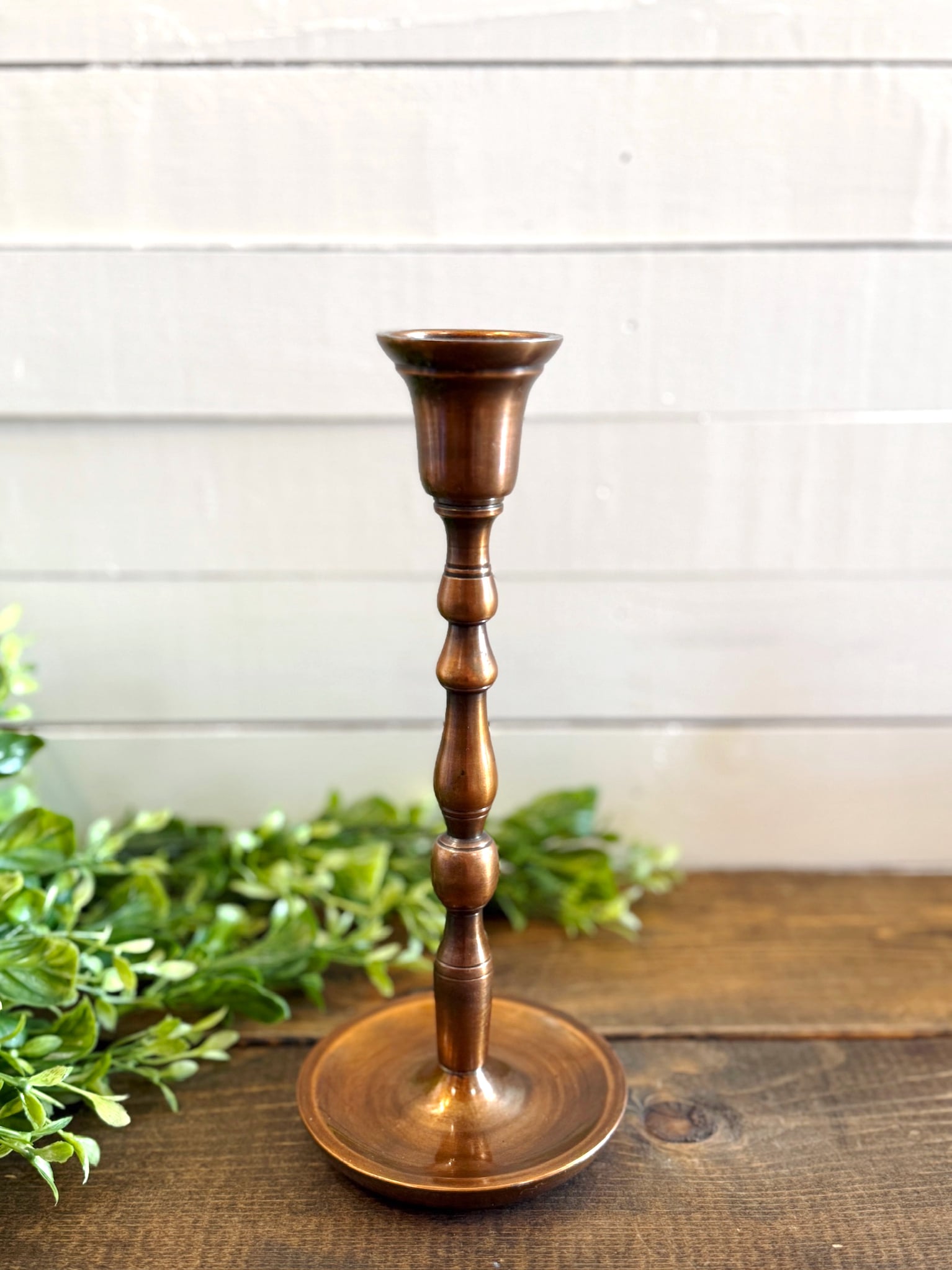 Antique copper medium taper candle holder with turned silhouette and round base, styled with greenery on a wooden surface.