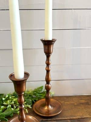 Small and medium antique copper candle holders with taper candles, styled on a wood surface with greenery.