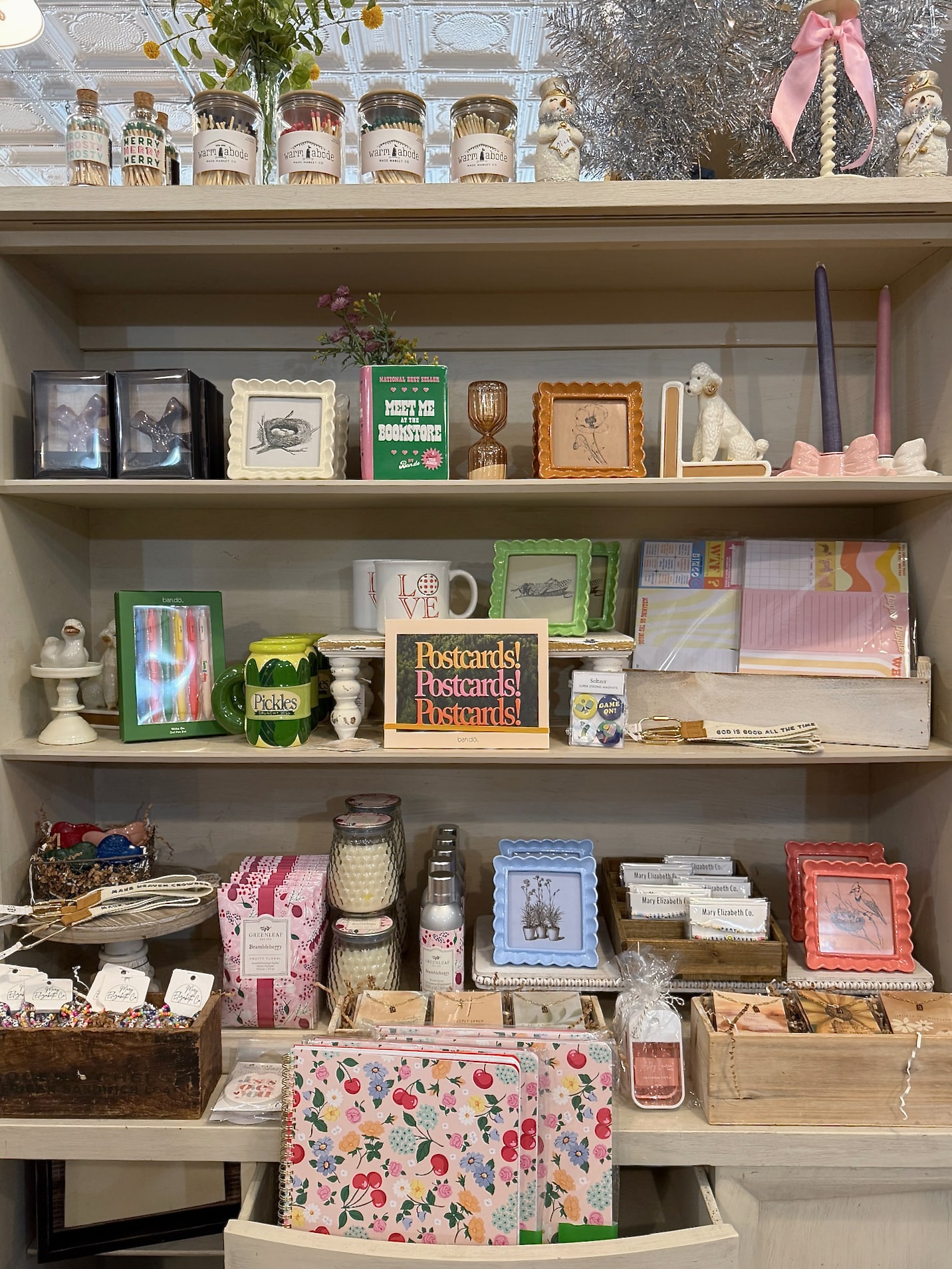 Shelf display featuring scalloped resin photo frames in pink, green, blue, coral, white, and brown alongside candles, notebooks, postcards, and seasonal decor inside Be Made Mercantile.