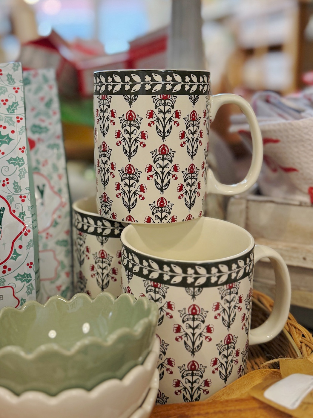 Cream stoneware mug with red and black floral block print and leafy rim border, displayed with holiday kitchen items.