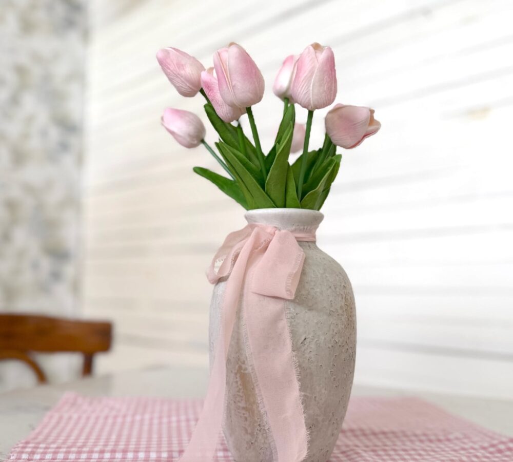 Medium Rustic White Vase filled with a Light Pink Tulip Bundle and tied with rose chiffon ribbon, displayed on a pink gingham square table runner.