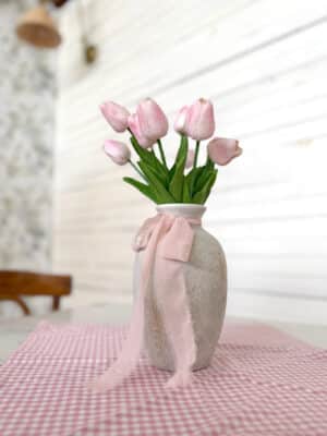 Medium Rustic White Vase filled with a Light Pink Tulip Bundle and tied with rose chiffon ribbon, displayed on a pink gingham square table runner.