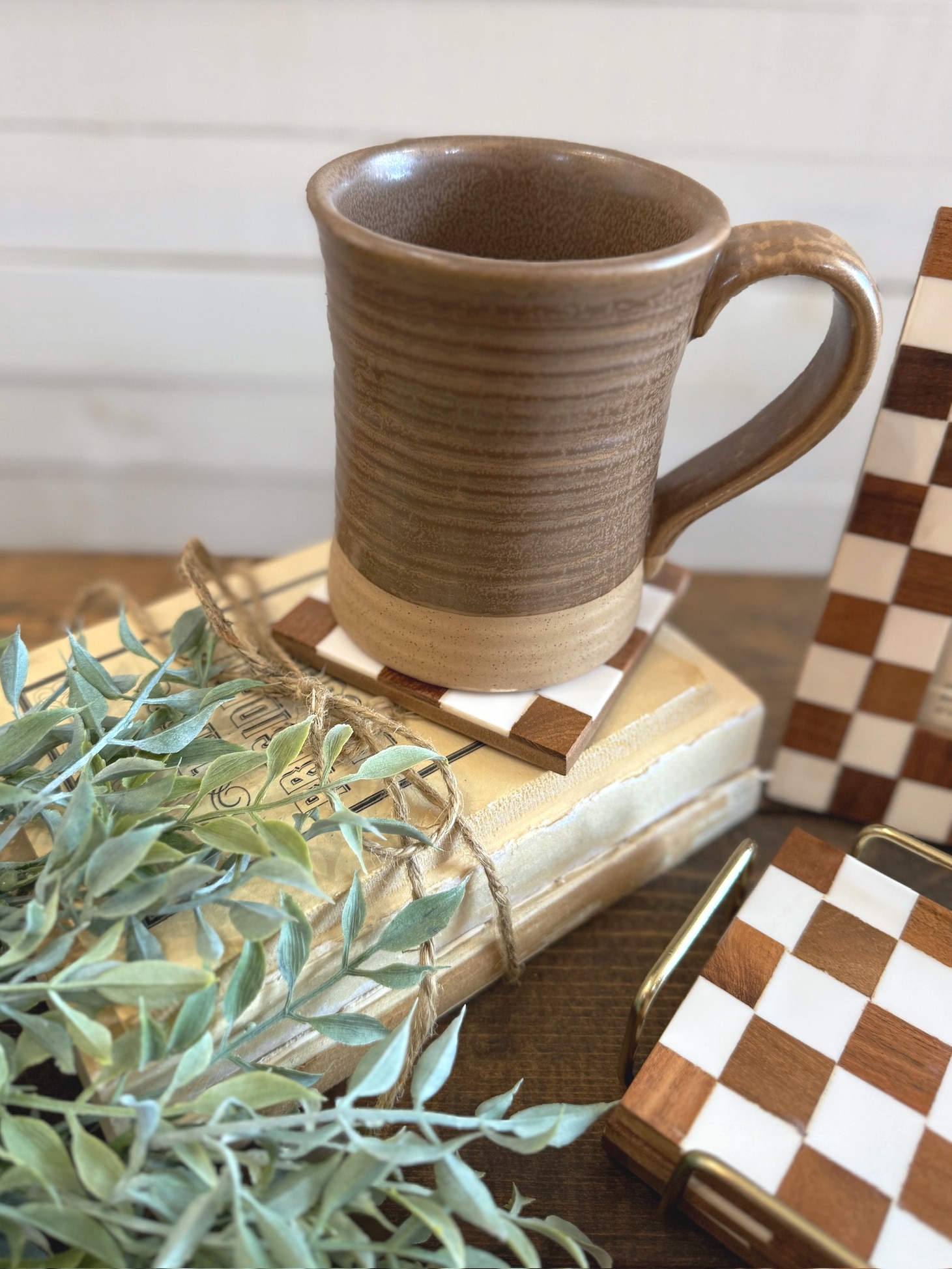 Brown glazed stoneware mug sitting on a checkered wood coaster, styled with vintage books, greenery, and matching decor.