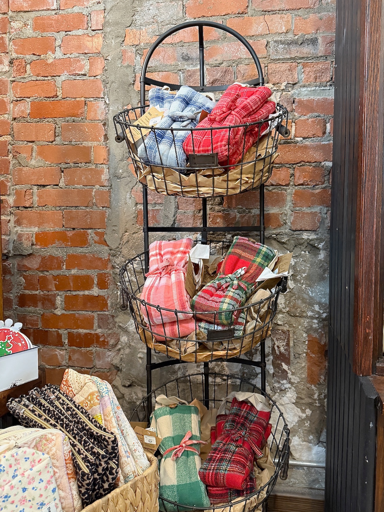 Display of Hot/Cold Neck Wraps in plaid and checkered flannel fabrics arranged in wire baskets against an exposed brick wall.