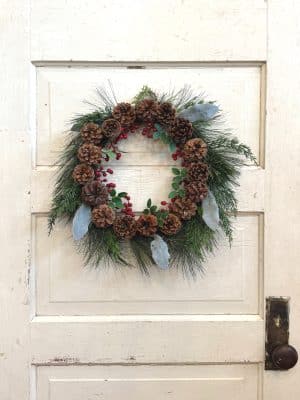 Christmas Pine Cone Berry Wreath hanging on a distressed white door, featuring pinecones, red berries, greenery, and soft velvet-like leaves.