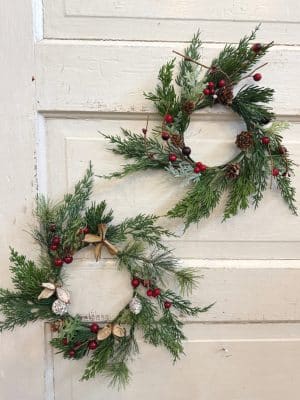 Two small holiday wreaths with greenery, red berries, and pinecones styled on a cream wooden door.