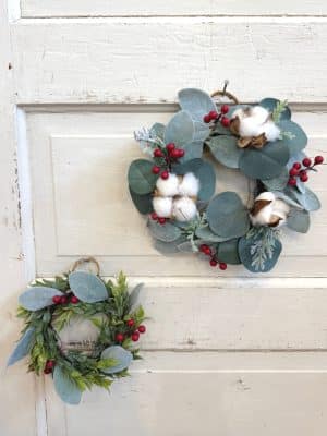 Two mini holiday wreaths with red berries, cotton accents, and greenery styled on a vintage white wood door.