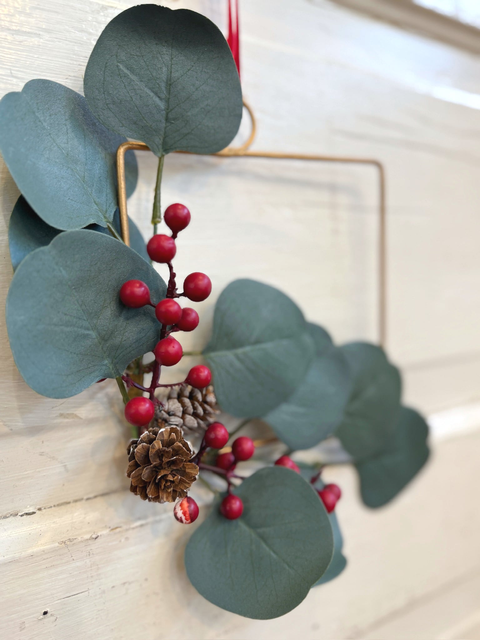 Close-up of faux eucalyptus, red berries, and pinecones on a gold square Christmas wreath.