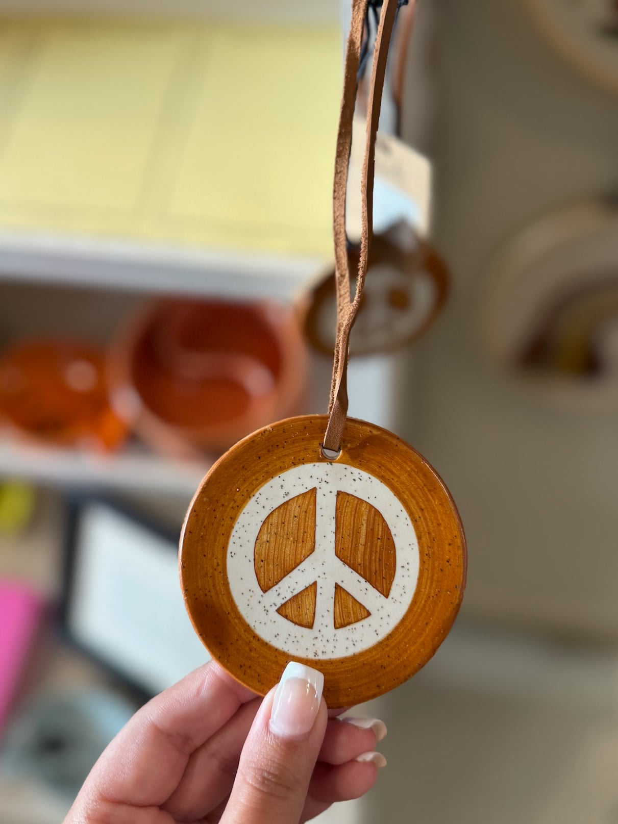 Brown clay peace sign ornament with a white raised symbol held in hand, featuring a leather hanging loop.