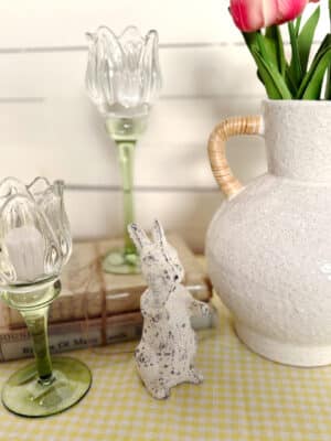 White Distressed Bunny Figurine styled with Tulip Glass Votive Candle Holders, a speckled vase with pink tulips, and stacked vintage books on a yellow gingham runner.