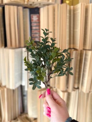 Hand holding a boxwood floral pick with lifelike foliage styled in front of neutral book decor.