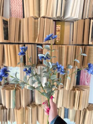 Faux blue blossom bush with vibrant clustered blooms and soft green leaves held against a neutral book-lined backdrop.