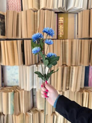Hand holding a faux blue mum spray with multiple textured blooms and natural-looking foliage.
