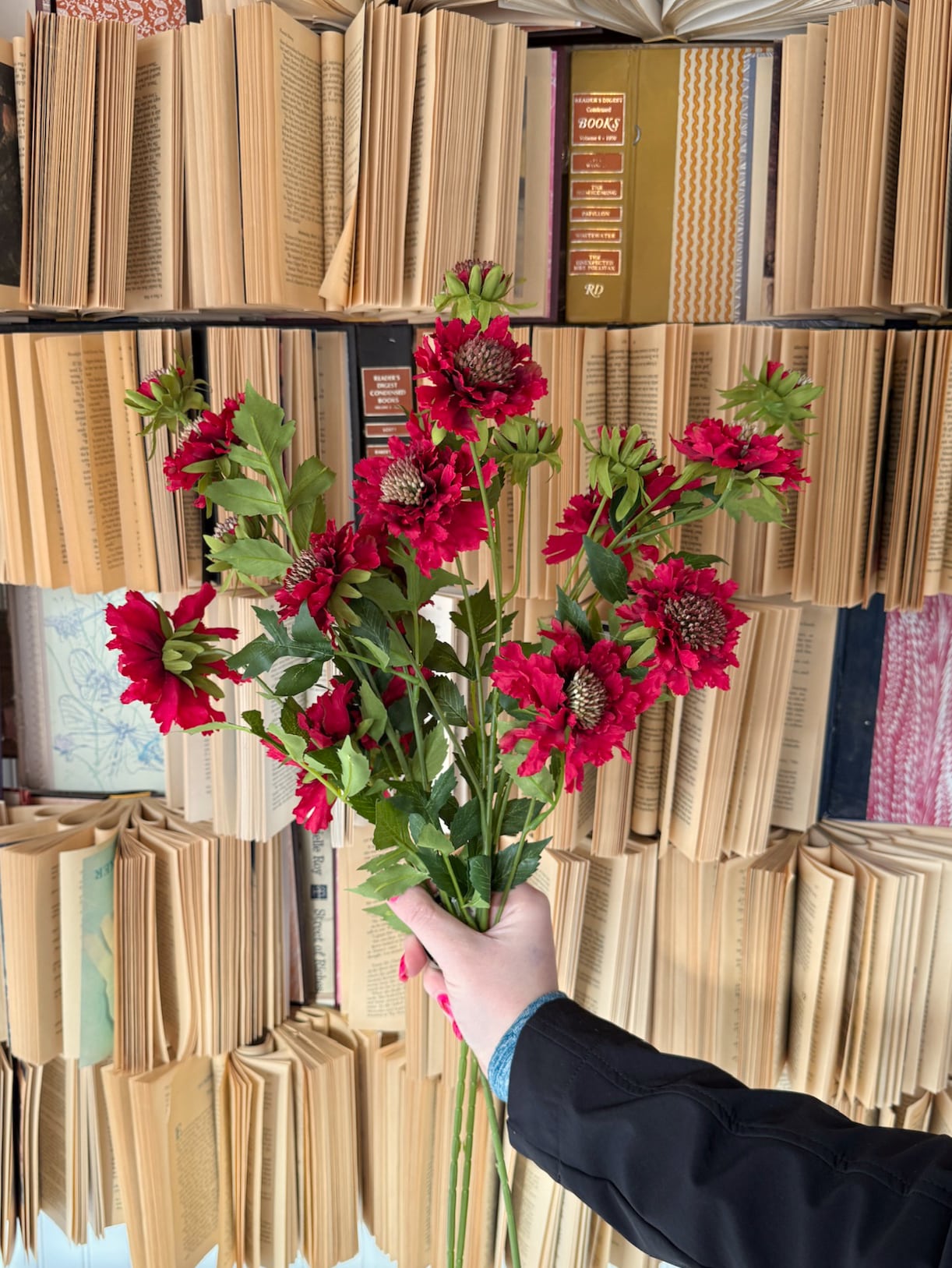 Hand holding a bouquet of lifelike red scabiosa stems with natural foliage.