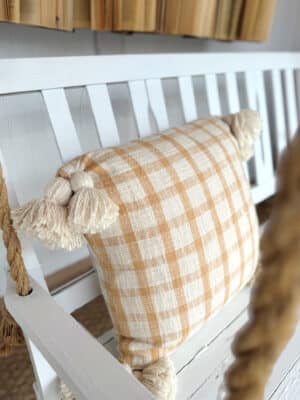Yellow plaid throw pillow with oversized tassels on a white bench.