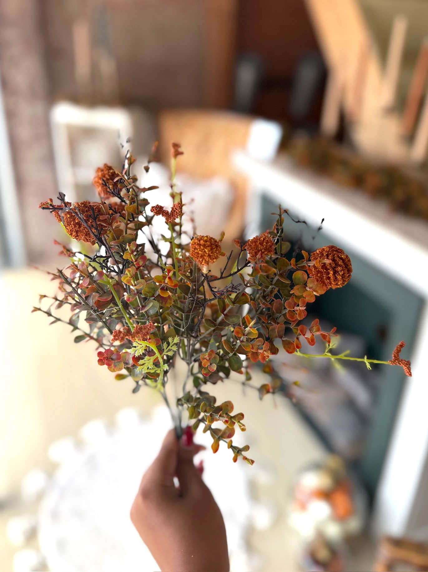 Hand holding a single Orange Pincushion Pick showing rust-orange blooms and soft greenery