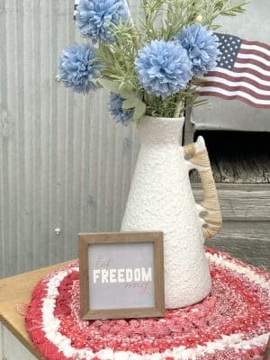 A small framed sign reading “Let Freedom Ring” sits on a red and white braided mat beside a tall off-white ceramic vase with rattan-wrapped handles and blue faux flowers. Two small American flags are tucked into the vase.