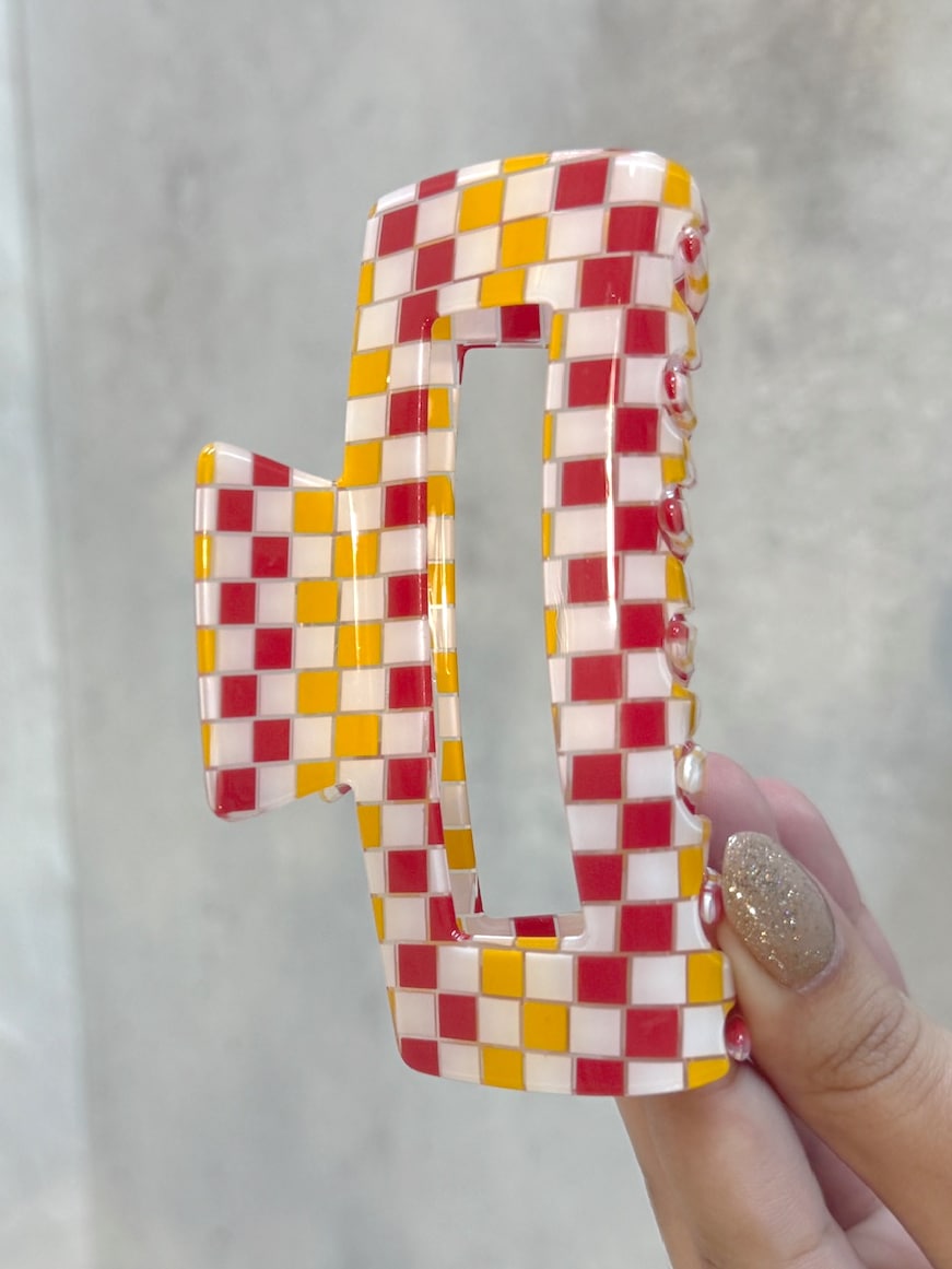 Hand holding a rectangular red, gold, and white checkered hair claw clip against a neutral background.